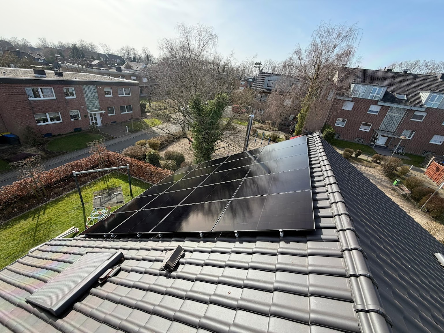 Frisch montierte Photovoltaikmodule auf einem dunklen Ziegeldach, Blick vom Dach über eine Wohnsiedlung